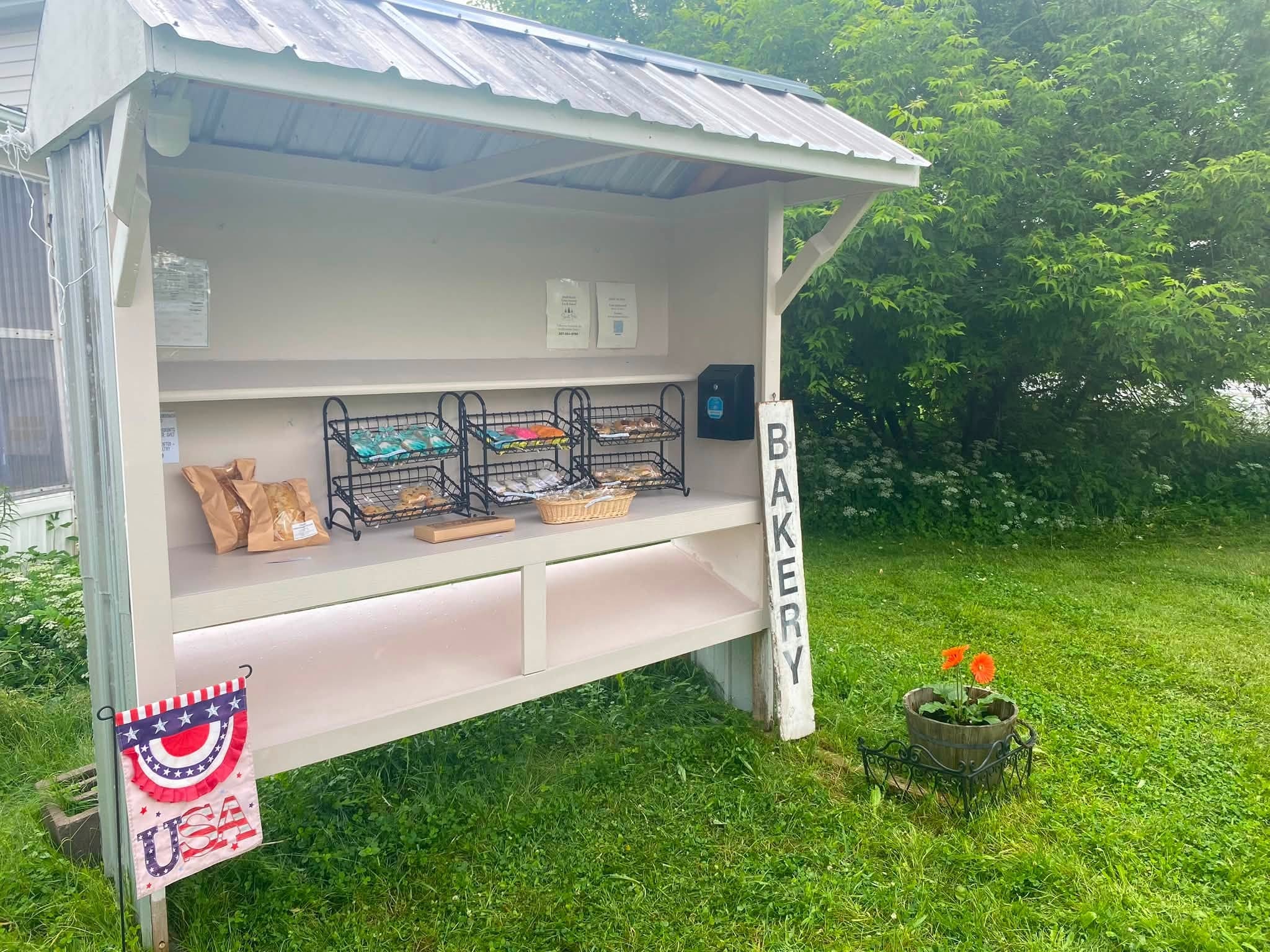 Sweet Pine Cookie Co. open-air white bakery stand with wire basket displays, USA bunting, and a potted orange flower in Bridgewater, Maine